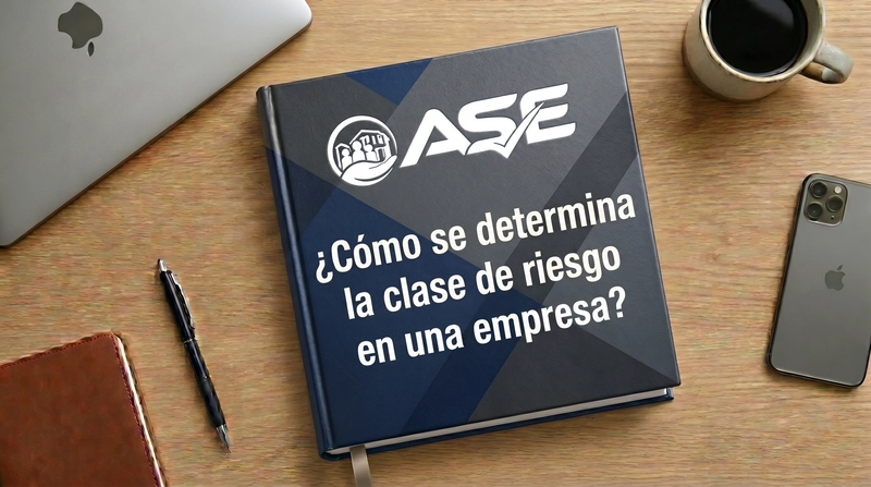 Libro azul marino con el logo de ASE y el texto Como se determina la clase de riesgo en una empresa en Colombia, sobre un escritorio de madera con laptop, celular, cuaderno y taza de cafe